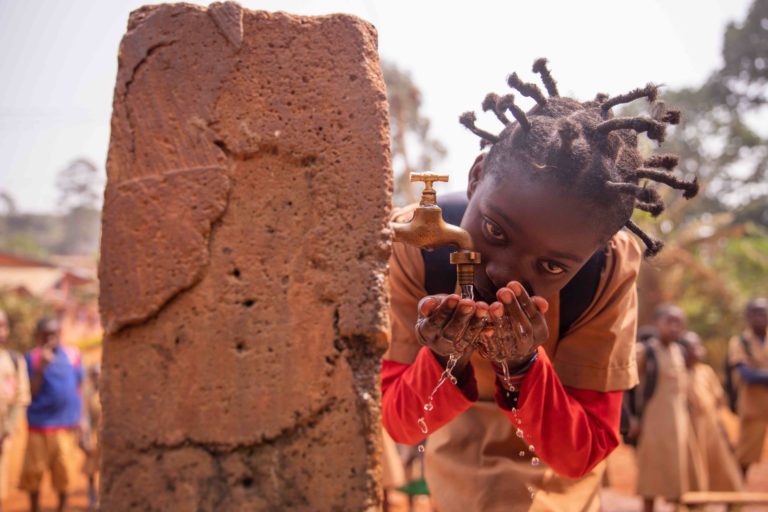 Fotografia di una bambina che si abbevera con le mani in una fontanella in un villaggio africano