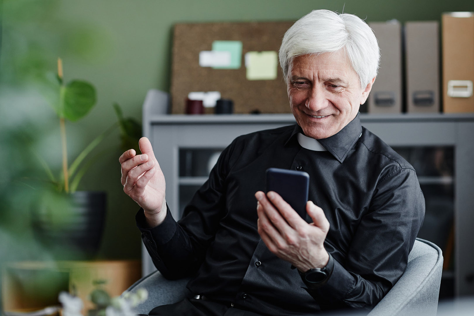 MAGISTER-CNEC-webinar-03-2025-1920×1080 Fotografia di un prete che guarda sorridente lo schermo del proprio smartphone